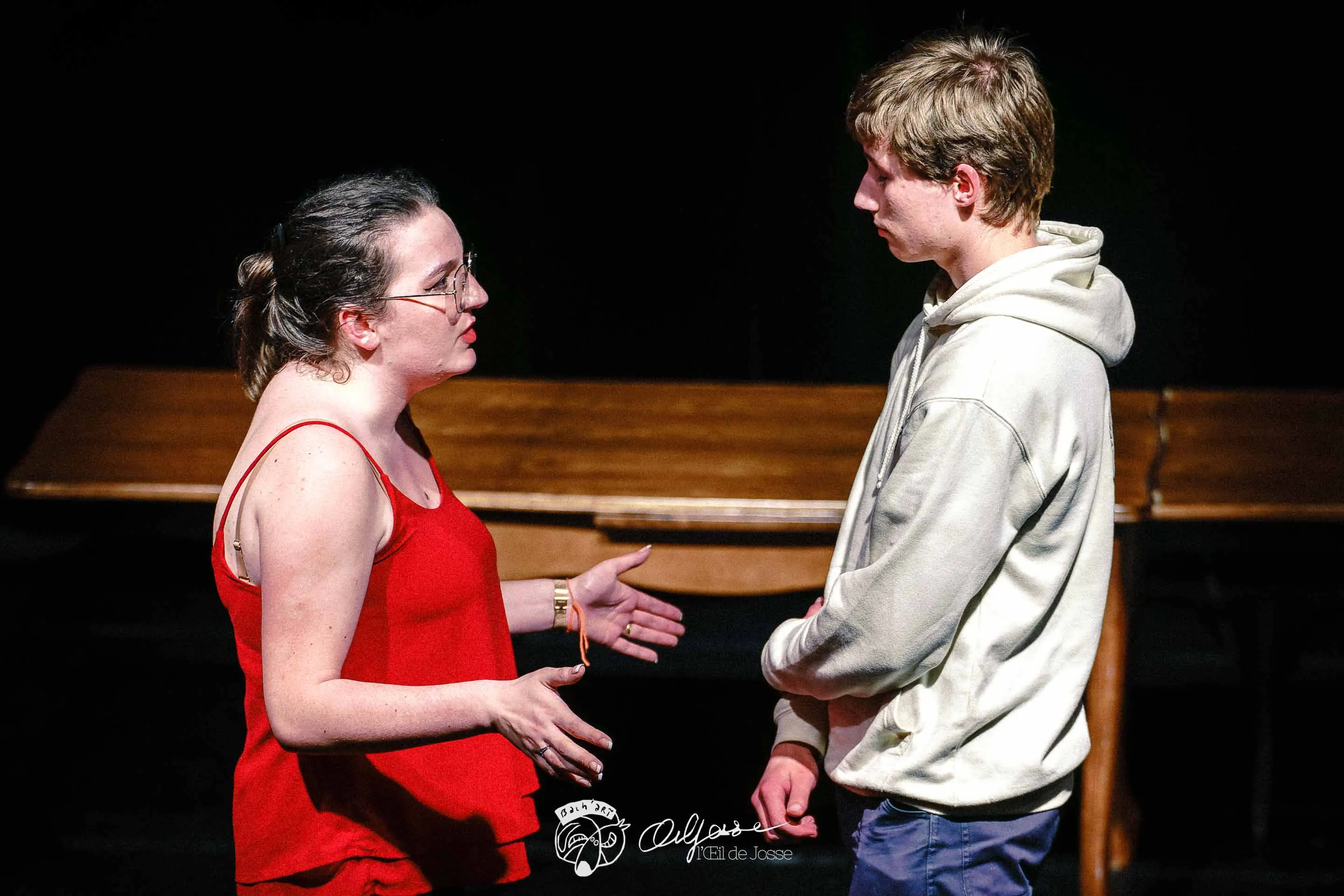 Une comédienne en rouge et un comédien en sweat se font face au-dessus d’une table.