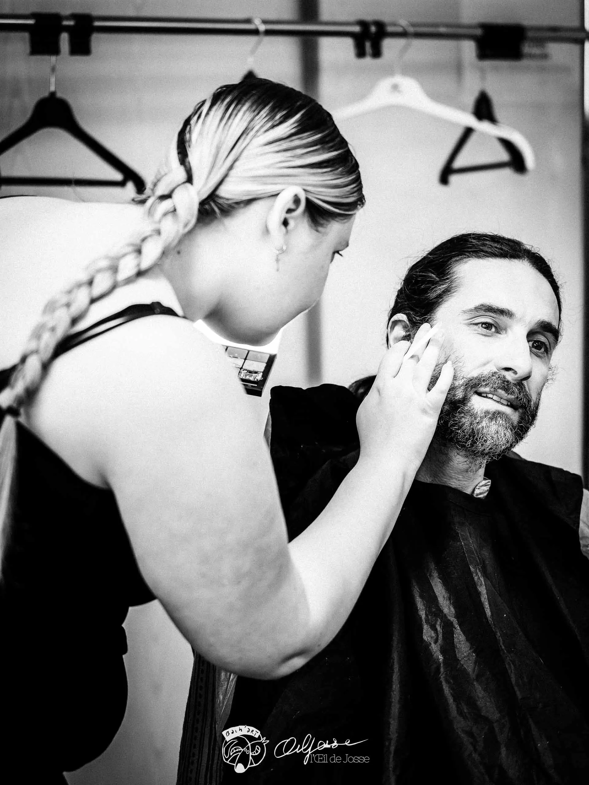 Une maquilleuse applique du fond de teint sur le visage d’un comédien barbu, en coulisses.